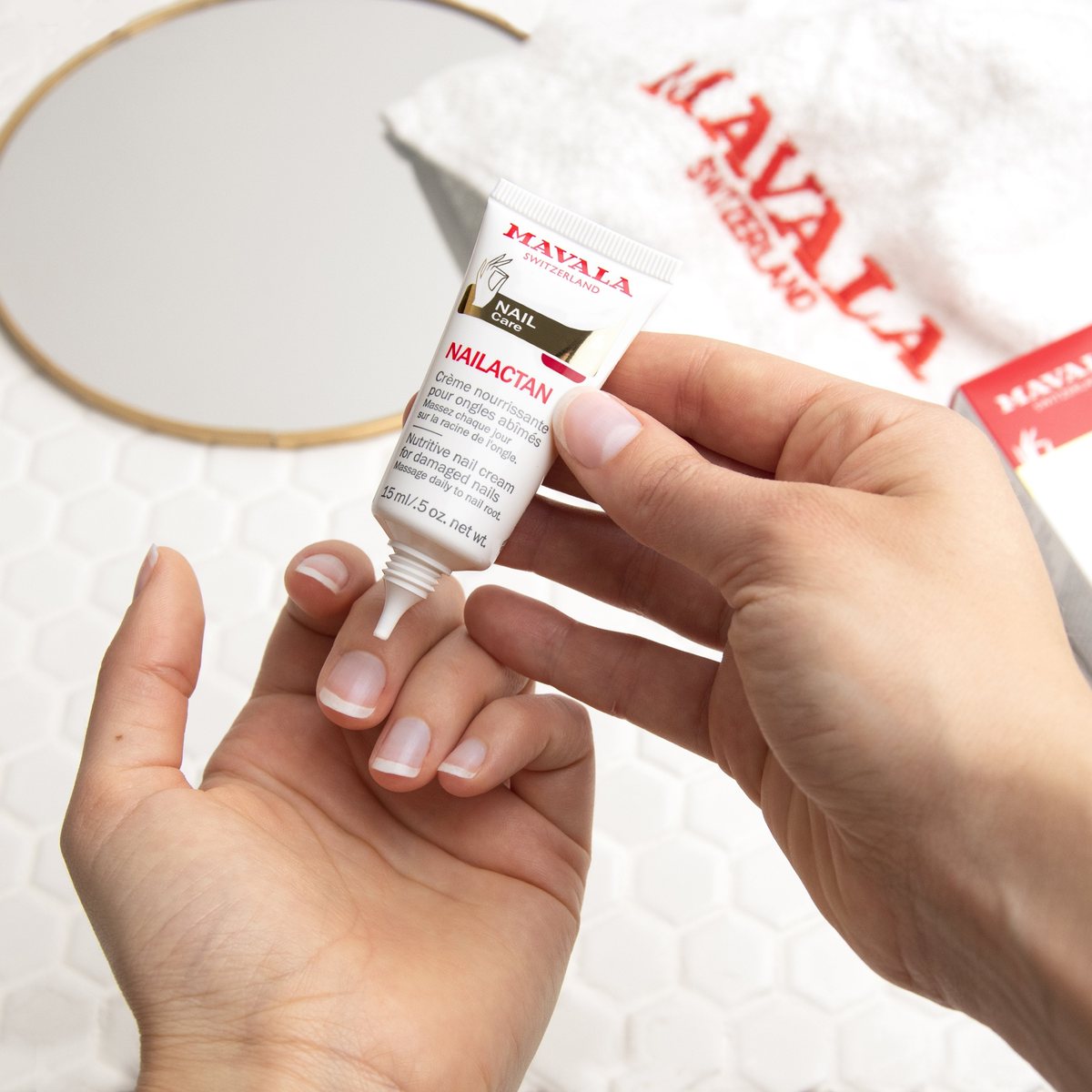 A 15 ml tube of MAVALA Nailactan nutritive nail cream being squeezed onto the base of a fingernail, with a MAVALA towel and a round mirror behind.