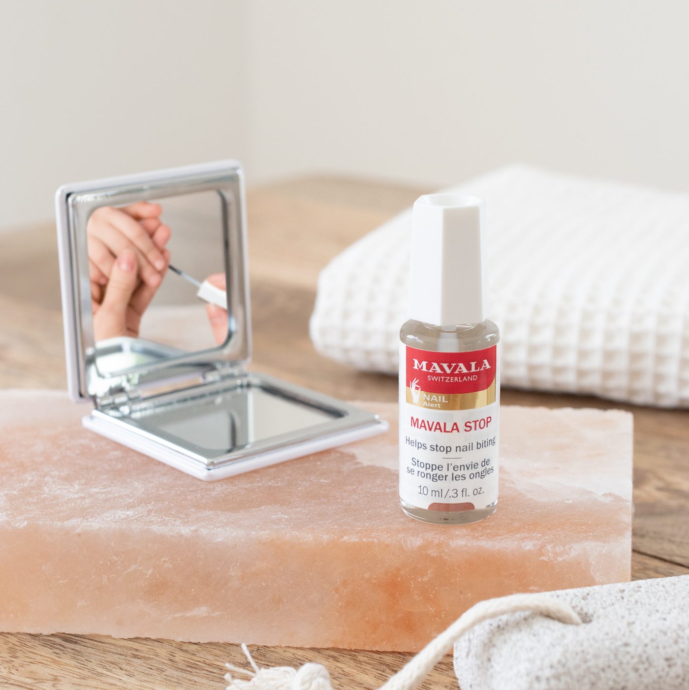 A bottle of Mavala Stop on a pink salt block, with a compact mirror reflecting a hand brushing the clear polish onto a nail.