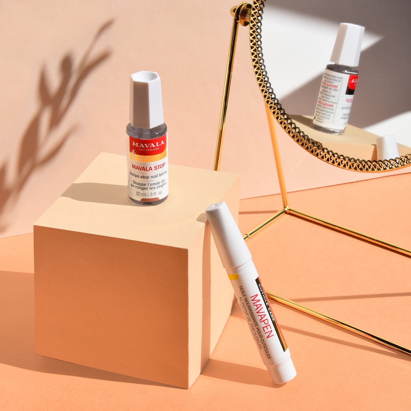 A bottle of Mavala Stop on a peach-coloured block beside a MAVAPEN cuticle oil pen, with a gold mirror behind.