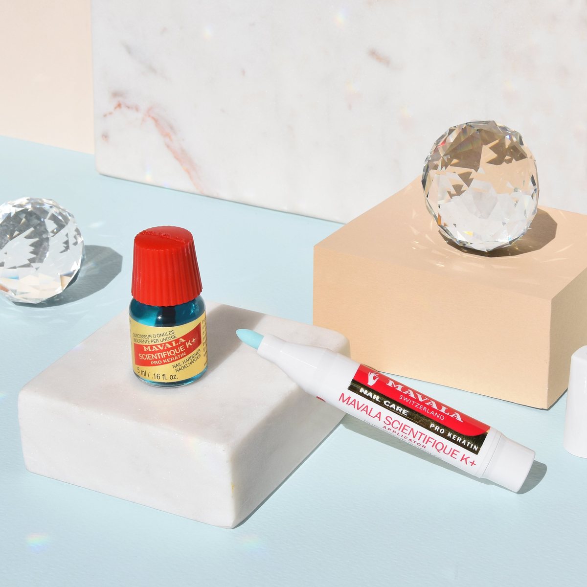 The 5 ml MAVALA Scientifique K+ nail hardener bottle on a marble block beside the MAVALA Scientifique K+ applicator pen, with cut-glass crystals on a pale blue background.